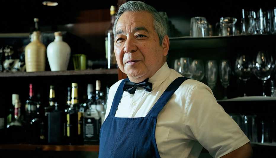 a bartender in an apron and a bowtie stands in front of bottles