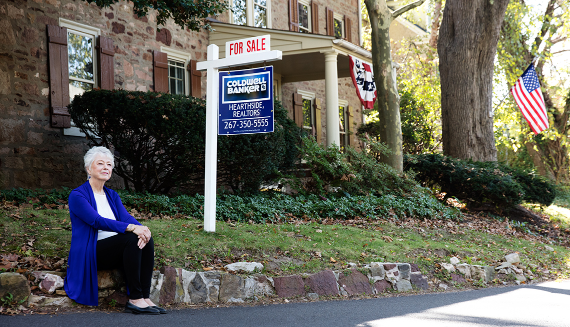 Real Estate Agent Joan Maguire sits on the curb in front of a house, near a For Sale sign