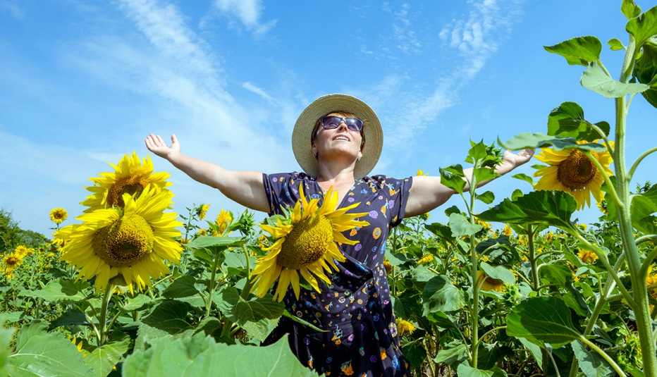 woman in sunglasses and straw hat, stands in field of sunflowers with arms outstretched to blue sky and sun