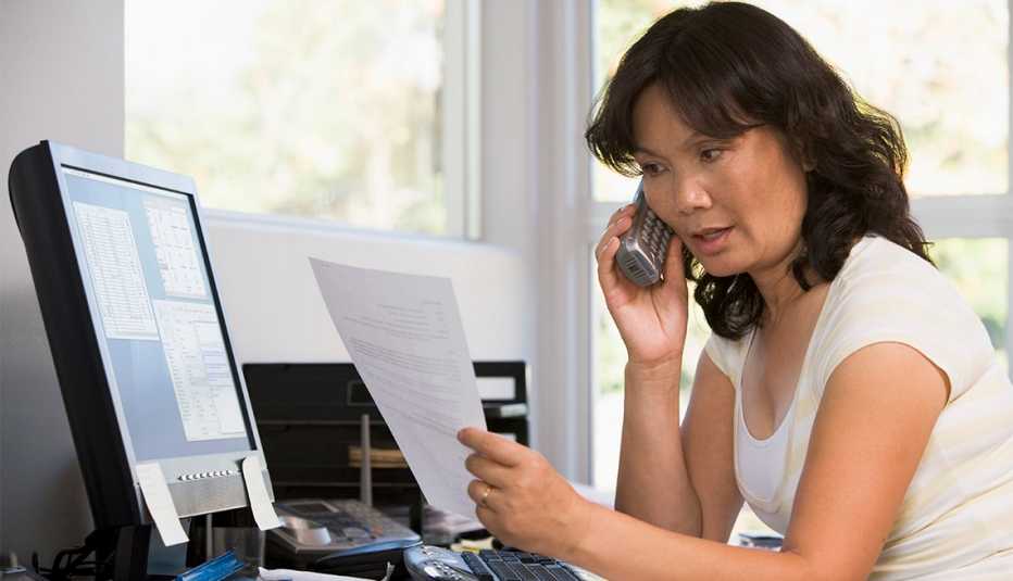 Mujer hablando por teléfono mientras revisa un documento y está sentada frente a la computadora.