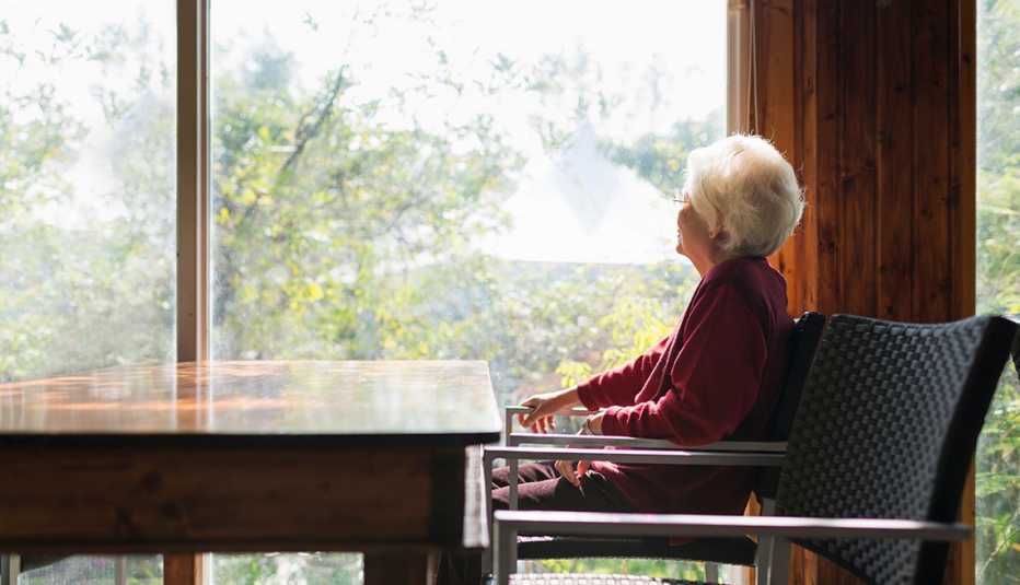 Mujer mayor sentada frente a una mesa mientras mira por la ventana.
