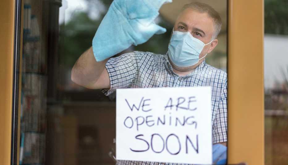 Hombre con tapaboca y guantes limpiando la puerta de entrada a un negocio con un letreo en inglés que dice abriremos pronto.