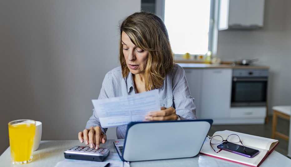 Mujer sentada en una mesa de la cocina haciendo cuentas con calculadora y computadora.