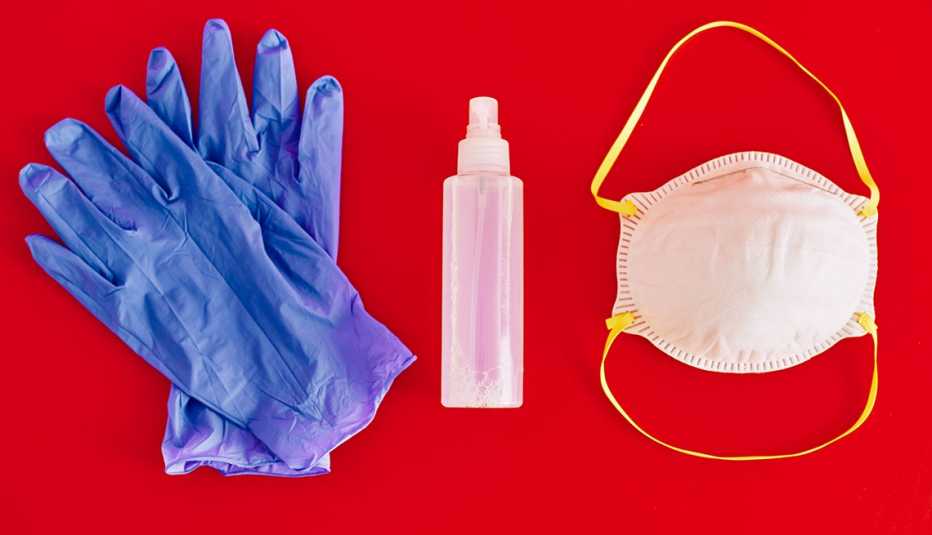 Guantes quirúrgicos, botella plástica con desinfectante de manos y una mascarilla sobre un fondo rojo.