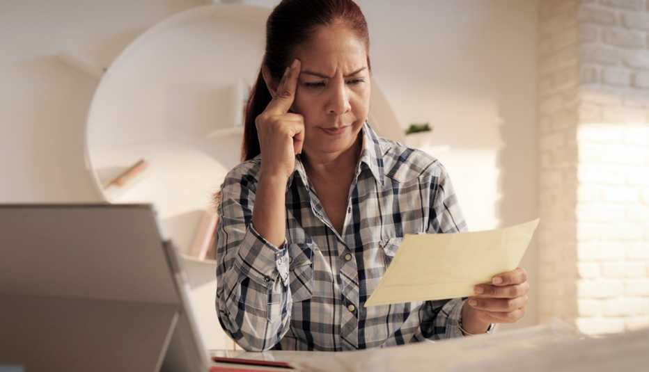 Mujer mira un documento mientras coloca el dedo índice en su cabeza para fijar su mirada. Al lado tiene una computadora.