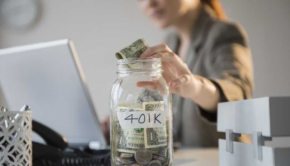 Mujer sentada en su escritorio y colocando dinero en una jarra marcada con 401k