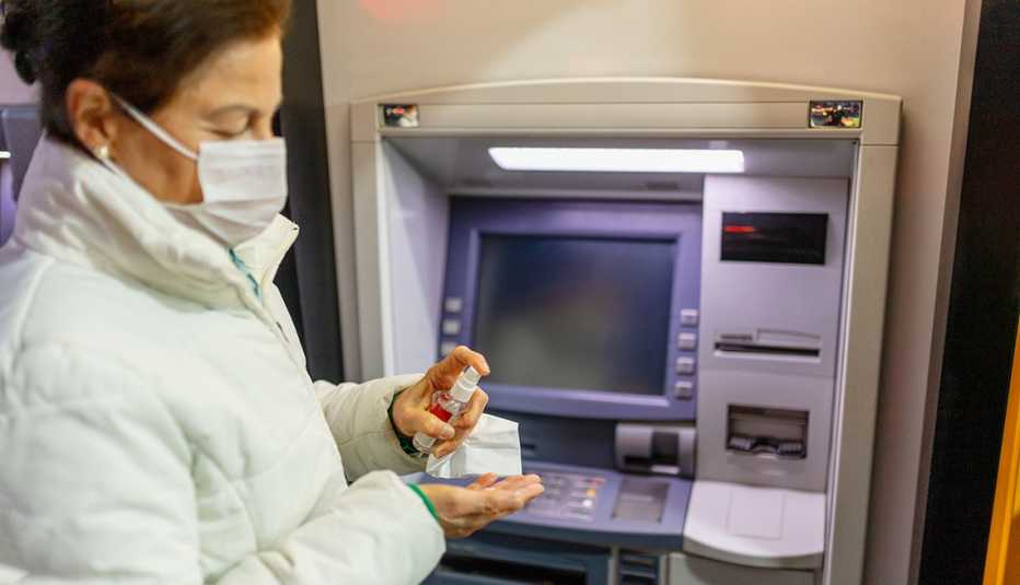 Mujer con una máscara en la boca frente a un cajero automático mientras se pone desinfectante en las manos.