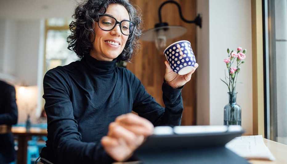 Mujer sonriendo mientras sostiene una tableta electrónica en una mano y una taza de café en la otra