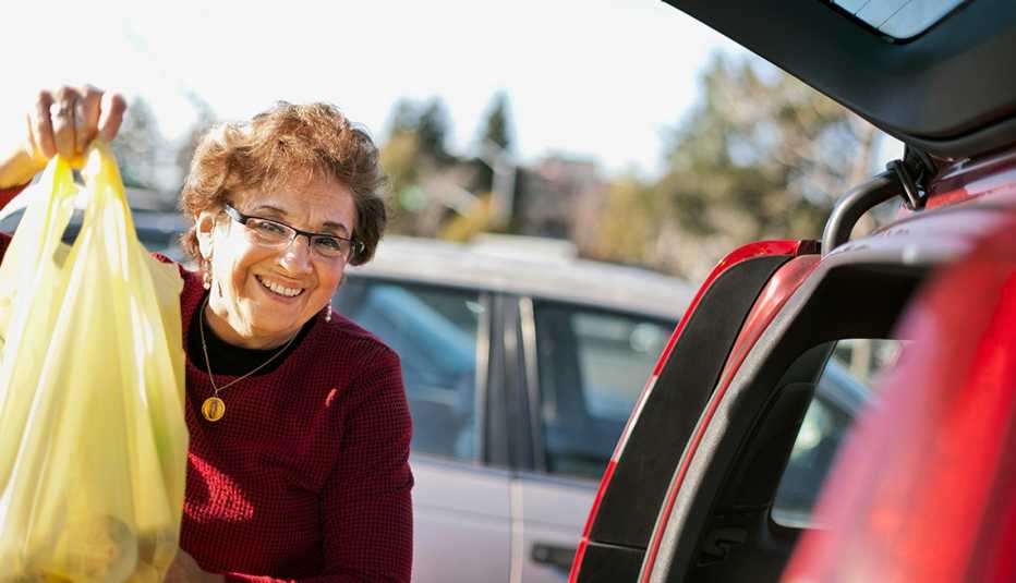Mujer mayor sonríe mientras sostiene una bolsa en un parqueadero y está al lado de su carro con la puerta del baúl abierta