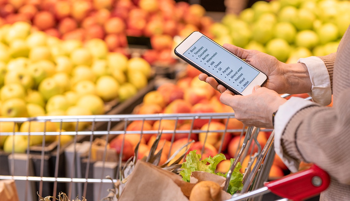 Manos de una mujer que revisa la lista de compras en su teléfono frente a la sección de frutas de un supermercado