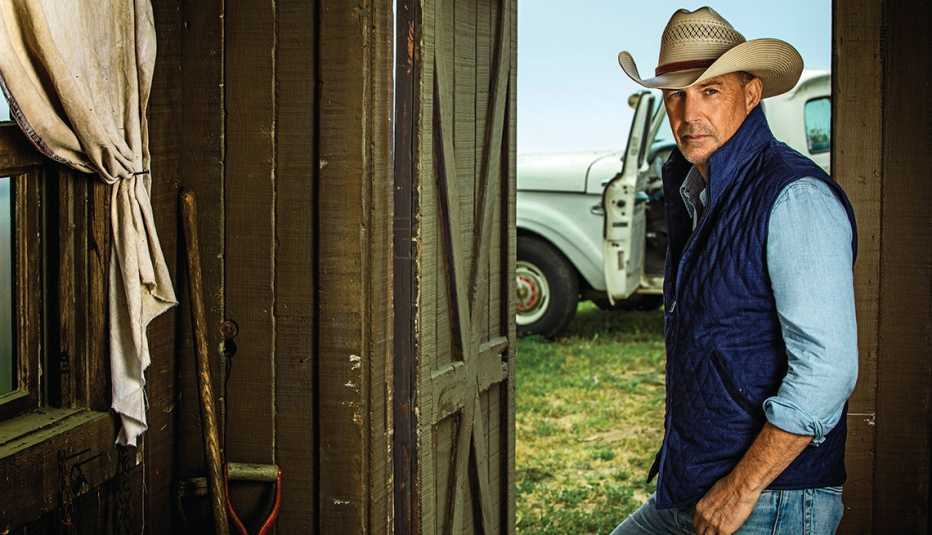 Kevin Costner en una casa de campo con sombrero, chaleco y una camioneta antigua al fondo.