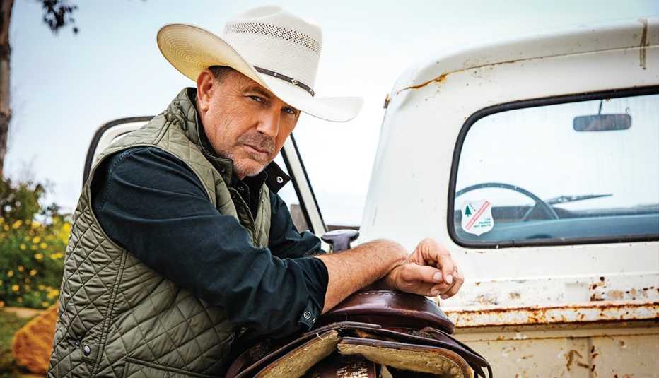 Retrato de Kevin Costner recostado sobre una silla de caballo en la parte trasera de una camioneta antigua.