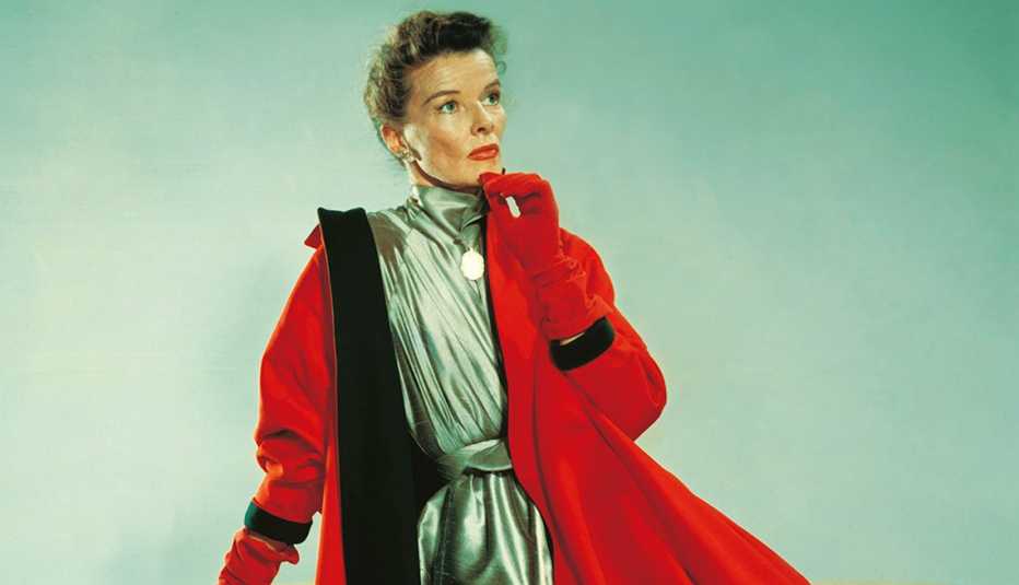 La actriz Katharine Hepburn posa con un vestido plateado y un abrigo rojo.