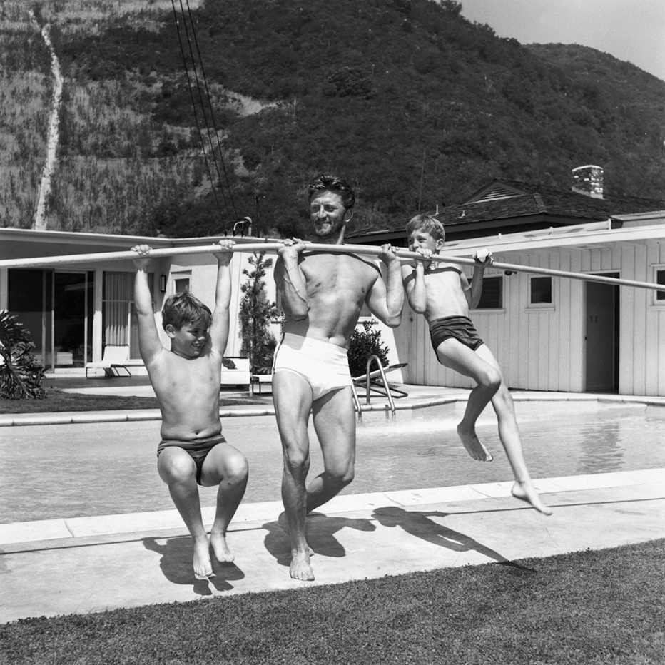 Foto en blanco y negro de los años 50 de Kirk Douglas levantando a sus hijos Michael y Joel con una vara frente a una piscina.