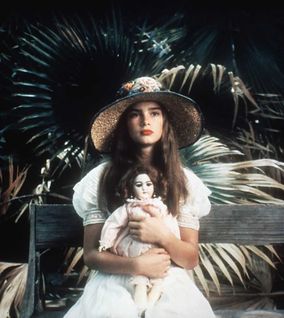 Brooke Shields en sus años adolescentes viste un sombrero, traje de verano y sostiene una muñeca en su falda