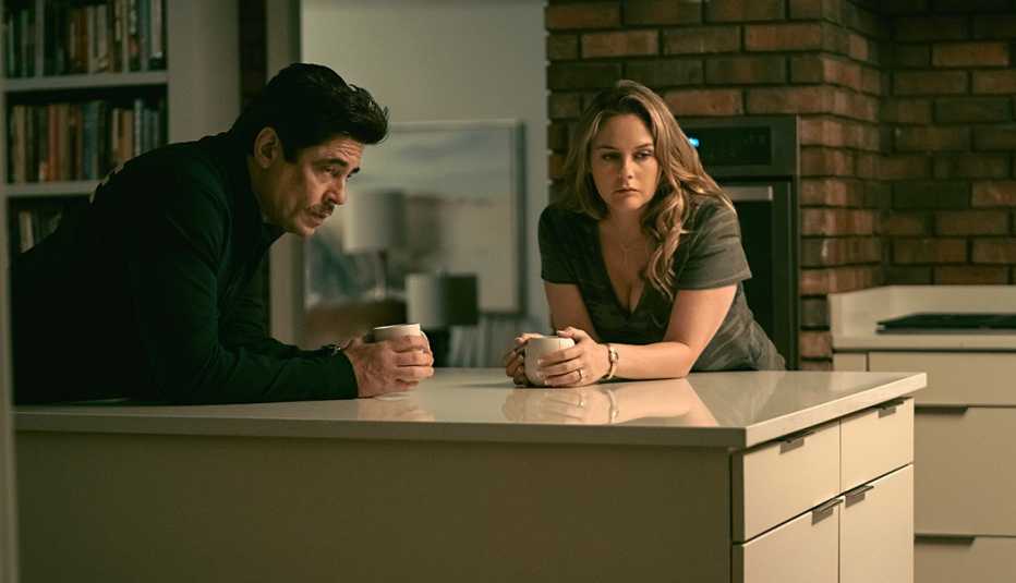 benicio del toro and alicia silverstone standing nearby each other at a kitchen island with coffee cups in a scene from the netflix film reptile