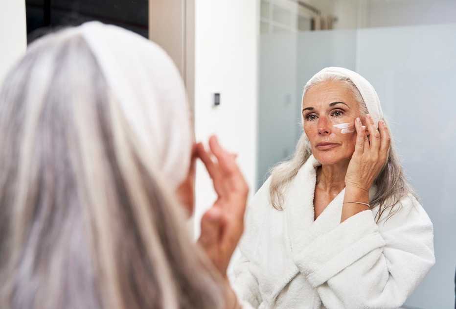 Una mujer aplicando crema en su cara frente a un espejo.