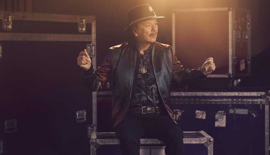 Carlos Santana sentado sobre cajas de equipo musical y tronando sus dedos en el aire.