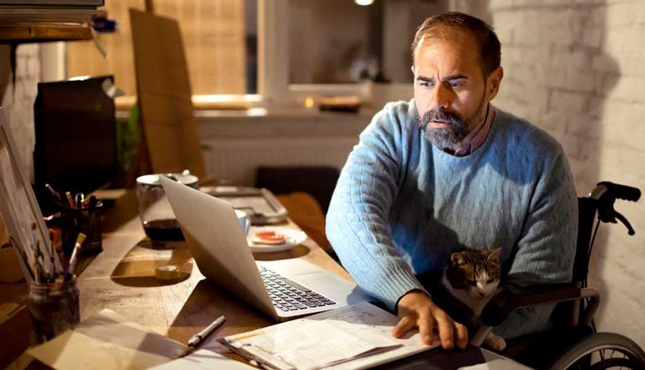 Hombre en silla de ruedas sentado en su escritorio en casa trabajando con su gato en las piernas