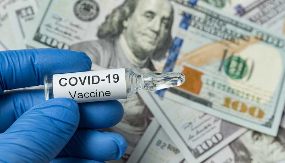 Mano con un guante sostiene una vacuna contra la Covid-19 y al fondo billetes de 100 dólares
