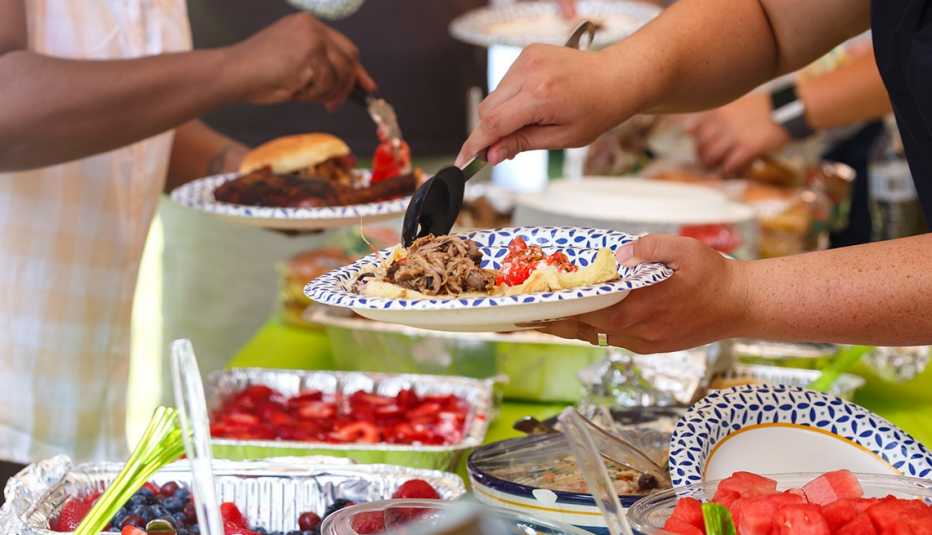 Invitados sirven platos con carne y fruta en una comida al aire libre informal de verano.