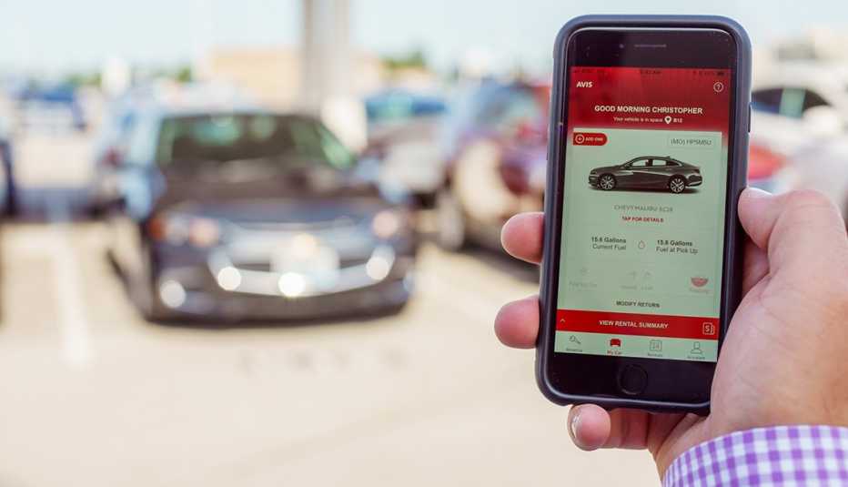 Hombre sostiene su teléfono con la aplicación de Avis en la pantalla frente a un estacionamiento de vehículos