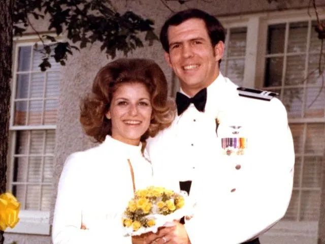 Tom y Yona McNish sonríen para una foto en su día de bodas. Ella tiene un vestido blanco de mangas largas y él viste un uniforme formal blanco de la Fuerza Aérea.