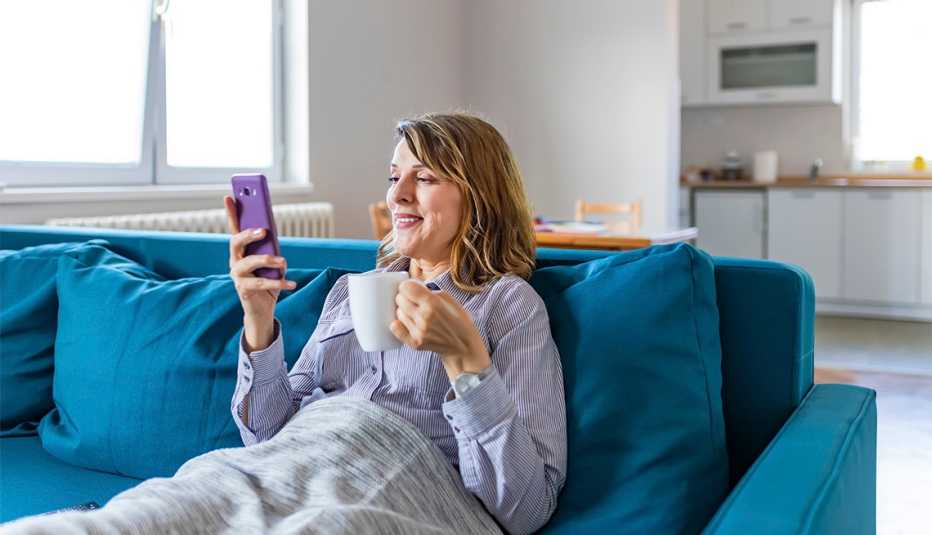 Mujer en un sofá con una tasa en una mano y un teléfono móvil en la otra.