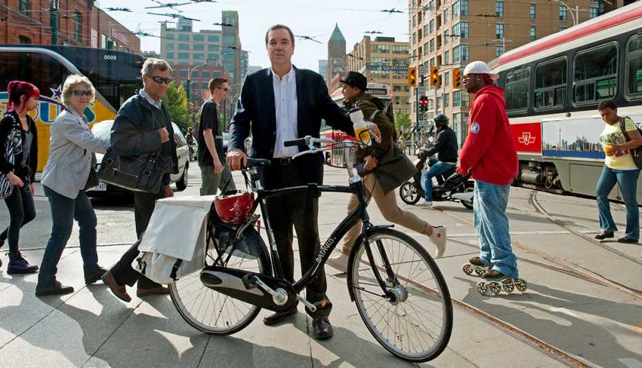 Gil Penalosa, Standing In Busy City Street Corner With Bicycle, Commuters And Buses In Background, Livable Communities, Why Older Adults Should Go Car-Free