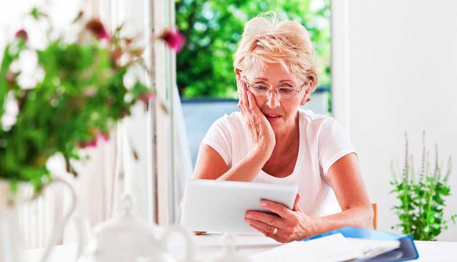 Mujer sentada en una mesa de su casa, con cara de preocupación y sosteniendo una tableta
