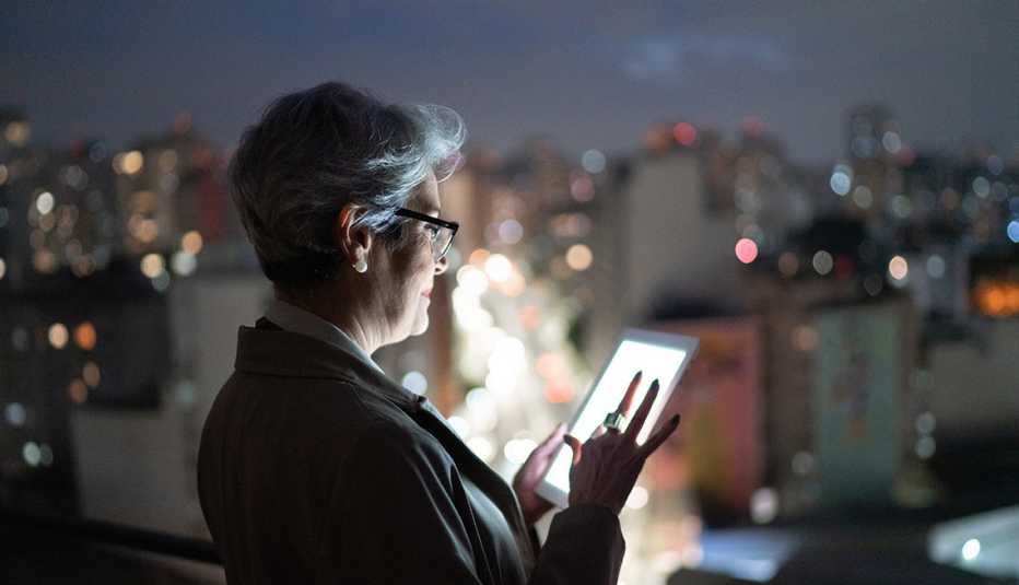 Mujer usando su tableta desde el techo de su casa y al fondo las luces de la ciudad