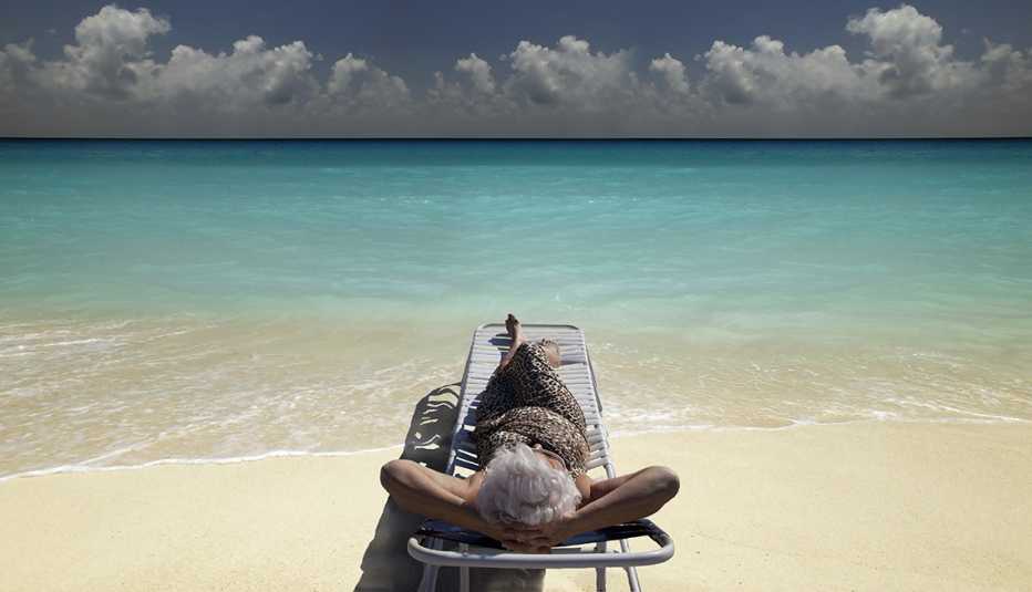 Mujer mayor recostada en una banca en la playa y mirando al cielo con el mar al fondo.