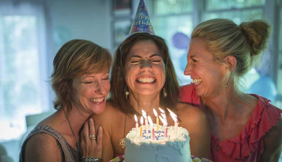 Mujer cierra los ojos, abre la boca y aprieta los dientes mientras sostiene un pastel con velas en medio de dos amigas