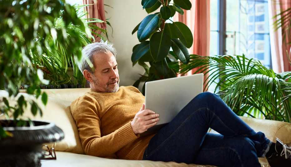 Un hombre con barba y cabello gris relajado en el sofá de su sala observando una computadora portátil