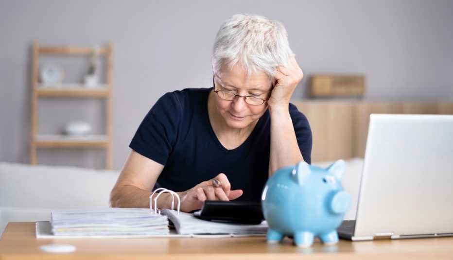 Mujer haciendo cuentas frente a su computadora, una alcancía azul, unas facturas y una calculadora