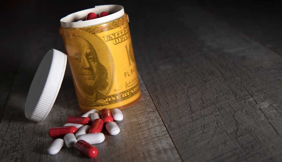 Cápsulas de medicina sobre una mesa al lado de un recipiente de pastillas envueltas en un billete de cien dólares