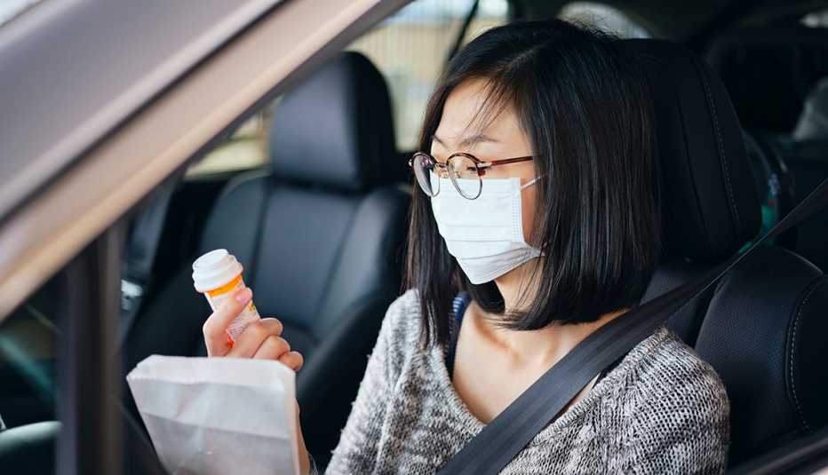 Una mujer con una máscara dentro de su auto mira un frasco de pastillas