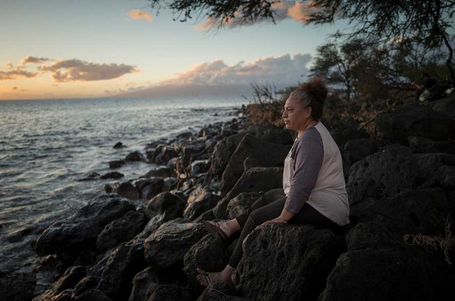 Etina Hingano sentada sobre rocas cerca del océano donde se agarró para sobrevivir el incendio forestal a principios de agosto del dos mil veinte y tres