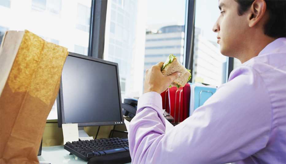 Hombre joven sentado en su oficina frente a un computador comiéndose un sándwich 