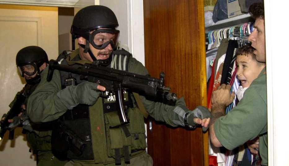 Militar armado apunta su arma a un niño y a un hombre que lo tiene en sus brazos.