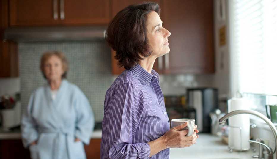 Una mujer pensativa toma café en su cocina, mientras una mujer mayor al fondo la observa.