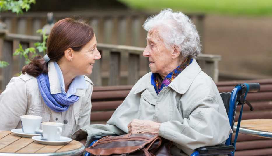 Una cuidadora conversa animadamente con una mujer mayor mientras toman café al aire libre.