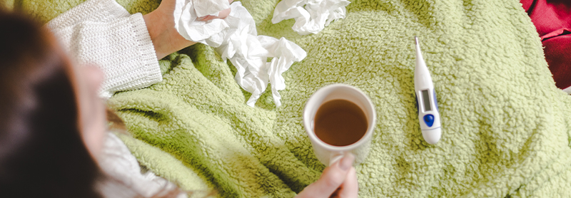 overhead shot of woman in bed holding cup of hot tea surrounded by thermometer and used tissues