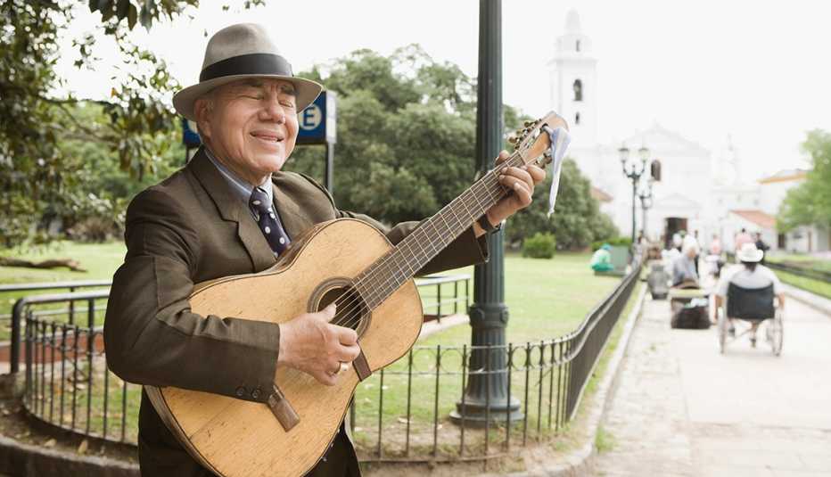 Hombre con sombrero y de traje con corbata toca la guitarra en una plaza pública. 