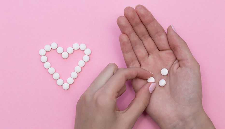Manos de una mujer sostienen unas pastillas, sobre un fondo rosa con pastillas formando un corazón