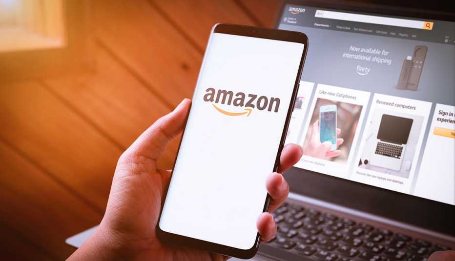 la mano de una mujer sostiene un teléfono inteligente con la aplicación de Amazon, y una computadora portátil en un escritorio con el sitio web de Amazon al fondo para comprar gotas que la FDA advirtió que no se compren