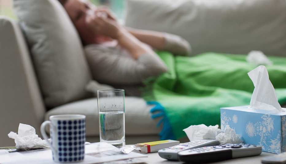 Al fondo una mujer enferma recostada en un sofá y sobre una mesa hay una taza, un vaso con agua y una caja de pañuelos desechables