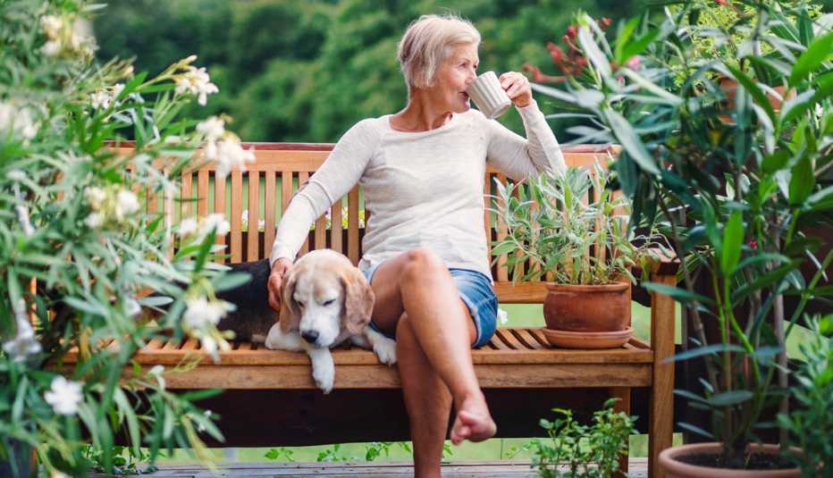 Una mujer sentada en un banco en un jardín tomando café con su perro.