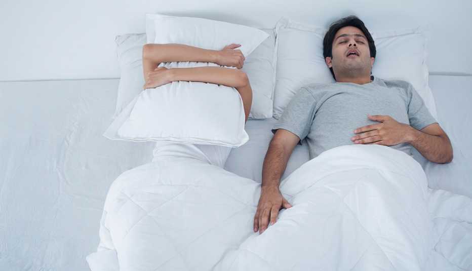 Pareja acostada en la cama, hombre dormido roncando y mujer cubriéndose con una almohada