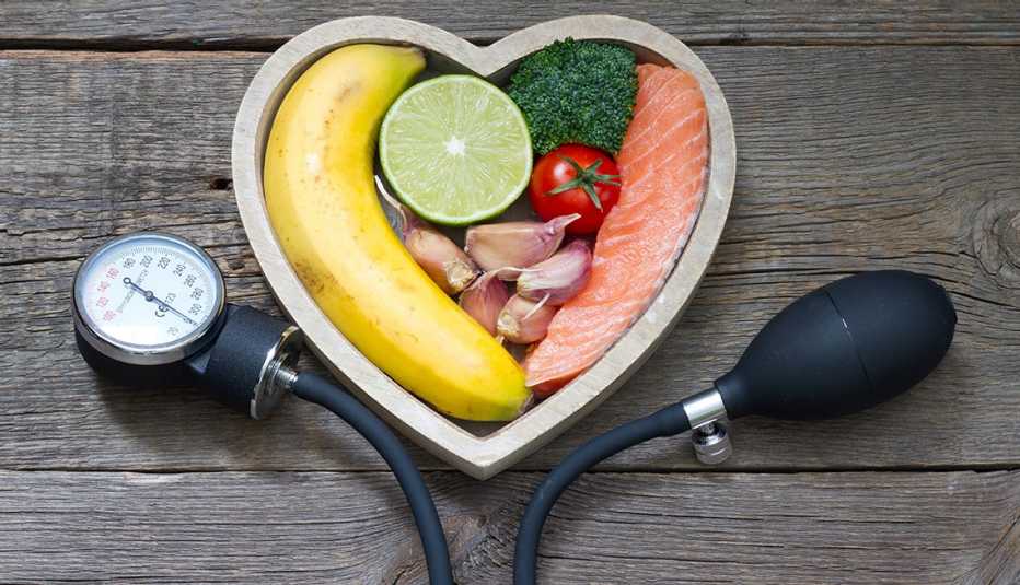 Un manguito de presión arterial aneroide enmarca una canasta en forma de corazón llena de alimentos saludables, incluidos productos agrícolas y salmón